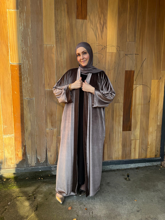 A woman wearing a long, flowing taupe-gray hijab and matching long robe stands in front of a wooden wall, holding the front of the robe closed.