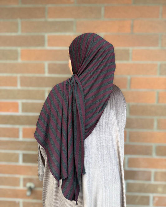 A person shown from the back wearing a dark hijab with diagonal stripes in black and deep red, paired with a light gray tunic, standing in front of a brick wall.