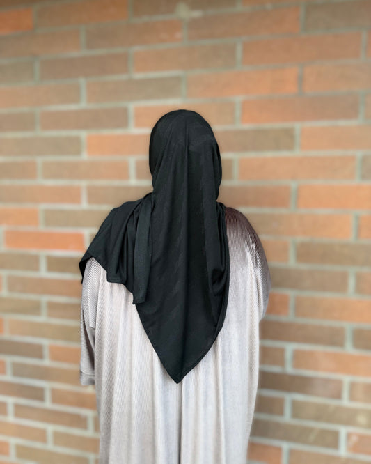 A person viewed from behind wearing a black full-head hijab and a light gray abaya, standing in front of a red brick wall.